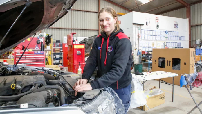 A young girl with dark blonde hair in slicked back works on a car with an open bonnet.