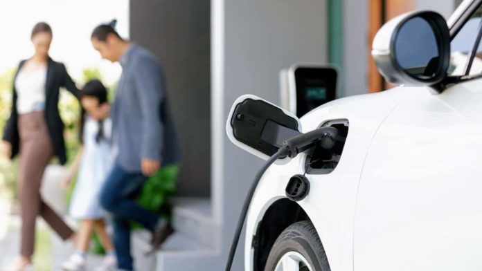 A cord attached to an electric vehicle charging it in the foreground and a blurred family in the background.