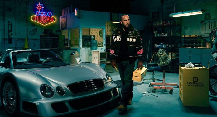 A man stands in a garage next to a silver sports car, holding a yellow folder. A neon sign reads "Gold Plush" in the background, reminiscent of the F1-Inspired Las Vegas Collection.